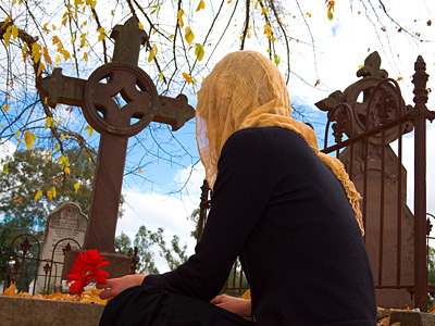Eine Person mit einem Kopftuch, die eine Blume hält, sitzt neben Grabkreuzen auf einem Friedhof. Herbstliche Blätter sind im Bild sichtbar.