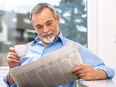 Ein älterer Mann sitzt am Fenster, liest eine Zeitung und hält eine Tasse in der Hand.