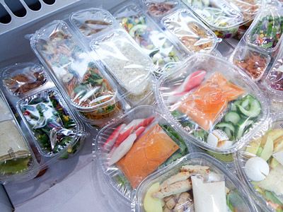 Verschiedene verpackte Lebensmittel in transparenten Behältern, darunter Salate, Gemüse und Fisch.