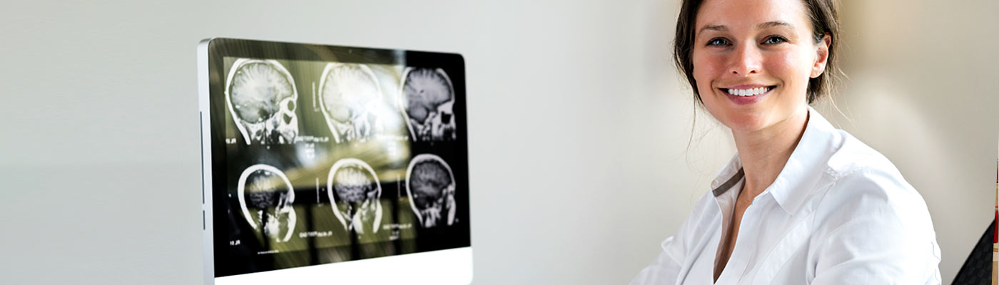 Eine lächelnde Frau in einem weißen Kittel sitzt vor einem Computerbildschirm, auf dem Gehirnscans angezeigt werden.