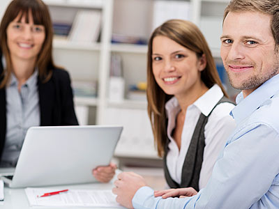 Drei Personen in einem Besprechungsraum, die lächeln und auf eine Kamera schauen. Auf dem Tisch befinden sich ein Laptop und Dokumente.