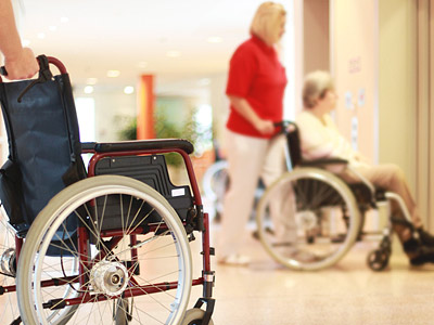 Ein Rollstuhl im Vordergrund, eine Person in rotem Shirt schiebt einen anderen Rollstuhl.