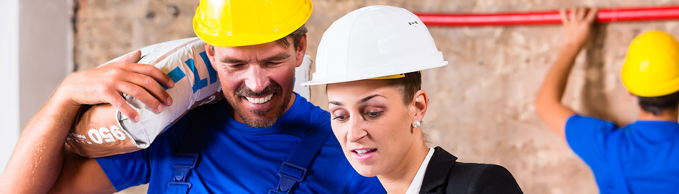 Ein Bauarbeiter mit gelbem Helm trägt einen Sack Zement und spricht mit einer Frau in Anzug, die einen weißen Helm trägt. Im Hintergrund ist ein weiterer Arbeiter zu sehen.