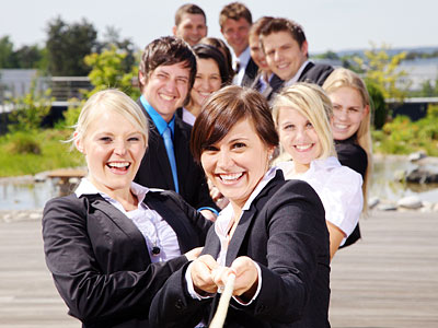 Eine Gruppe von fröhlichen Menschen in Geschäftskleidung, die an einem Seil ziehen, vor einer grünen Landschaft.