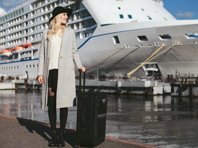 Eine Frau mit Koffer steht vor einem Kreuzfahrtschiff. Sie trägt einen langen grauen Mantel und einen schwarzen Hut.
