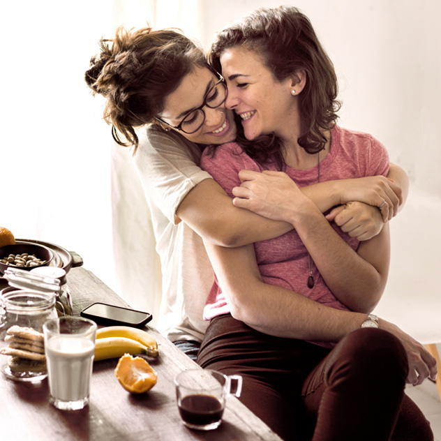 Zwei Frauen sitzen an einem Tisch, umarmen sich und lächeln sich an. Auf dem Tisch stehen Snacks und Getränke.