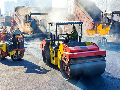 Bauarbeiten mit Straßenwalzen und anderen Maschinen. Rauch und Arbeiter sind sichtbar.