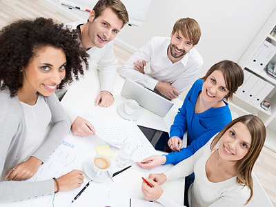 Fünf Personen sitzen an einem Tisch, lächeln und diskutieren über Dokumente. Tassen und Tablets sind ebenfalls sichtbar.