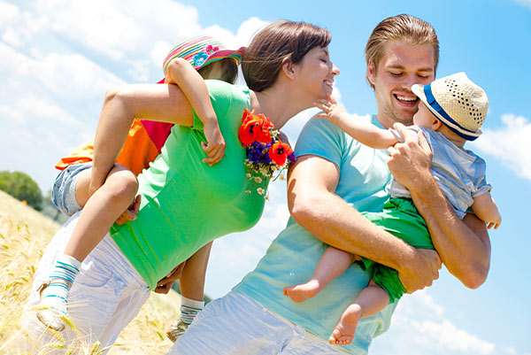 Eine fröhliche Familie im Freien, die in einem Feld steht. Die Eltern tragen jeweils ein Kind auf dem Rücken. Es ist sonnig und die Familie lächelt.