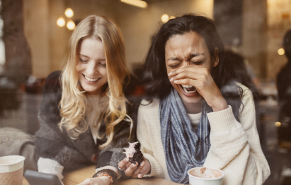 Zwei Frauen sitzen in einem Café und lachen. Eine von ihnen hält ein Dessert in der Hand.