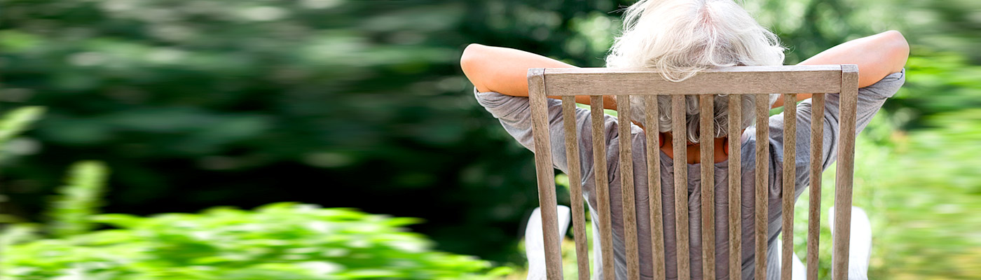 Eine Person mit grauen Haaren sitzt entspannt auf einem Stuhl im Freien, umgeben von grüner Natur.