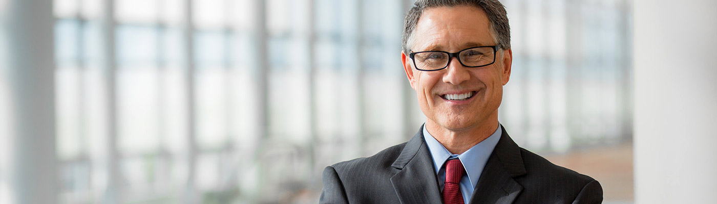 Ein lächelnder Mann in Anzug und Krawatte steht vor einem hellen, modernen Hintergrund.