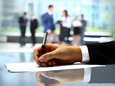 Eine Hand hält einen Stift und schreibt auf ein Blatt Papier, im Hintergrund sind Menschen in einem Geschäftsumfeld zu sehen.