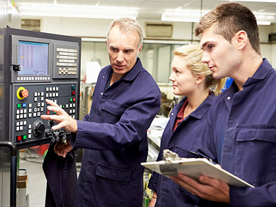 Ein Trainer erklärt Bedienfunktionen an einer Maschine, während zwei Auszubildende aufmerksam zuhören.