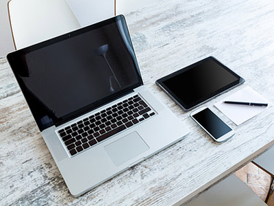 Ein Laptop, ein Tablet, ein Smartphone und ein Notizblock mit einem Stift auf einem Holztisch.