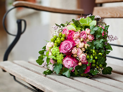 Ein bunter Blumenstrauß mit rosa Rosen, kleinen weißen und grünen Blumen, umgeben von Blättern, liegt auf einer Holzbank.
