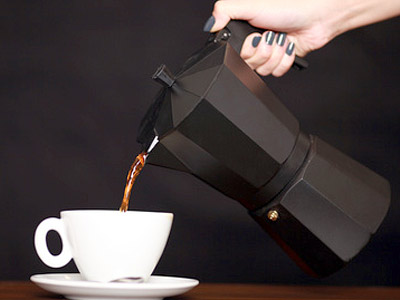 Eine Person gießt Kaffee aus einer schwarzen Moka-Kanne in eine weiße Tasse.