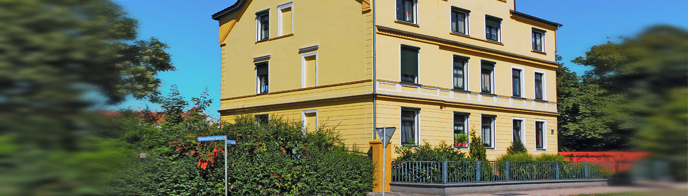 Ein gelbes Gebäude mit mehreren Fenstern, umgeben von Bäumen und Sträuchern, in einem klaren blauen Himmel.