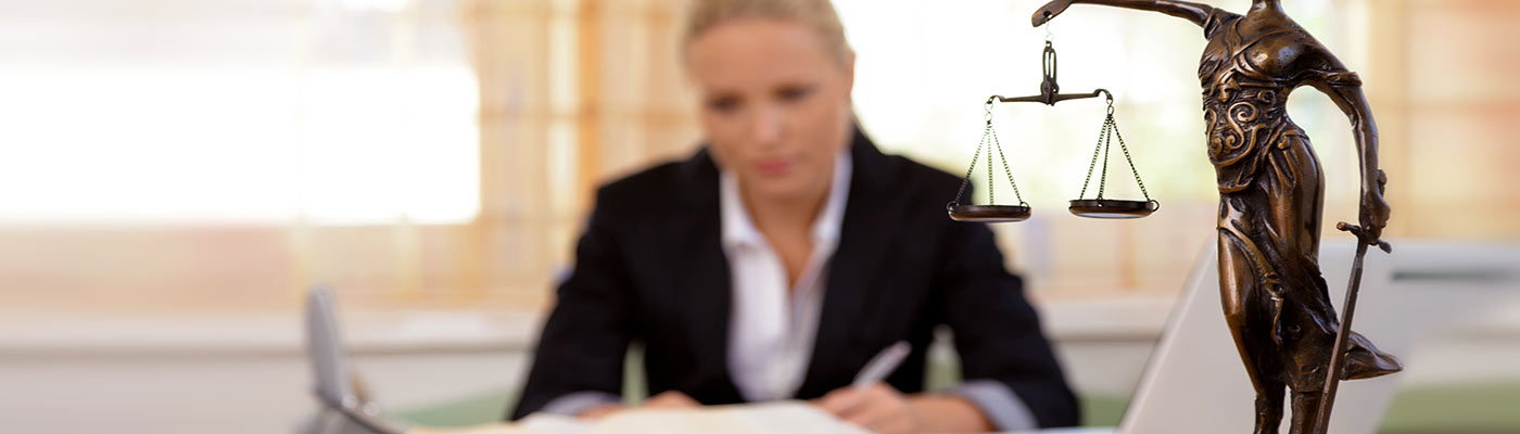 Eine Anwältin sitzt am Schreibtisch, während eine Statue der Justitia mit einer Waage im Vordergrund sichtbar ist.