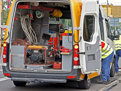Ein Lieferwagen mit offenem Heck, der mit Werkzeugen und Ausrüstungen für Reparaturarbeiten beladen ist. Zwei Personen in Warnwesten sind in der Nähe.