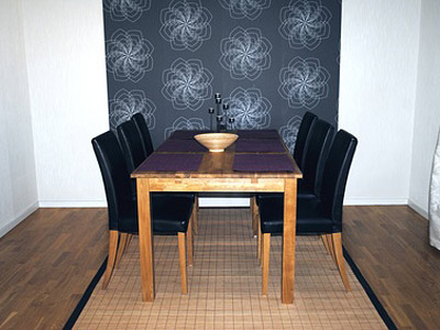 Ein Esszimmer mit einem Holztisch, sechs schwarzen Stühlen und einer dekorativen Schüssel auf dem Tisch. Im Hintergrund befindet sich eine blumige Tapete.