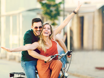 Ein lächelndes Paar fährt fröhlich auf einem Fahrrad, die Frau hat die Arme in die Luft gestreckt.