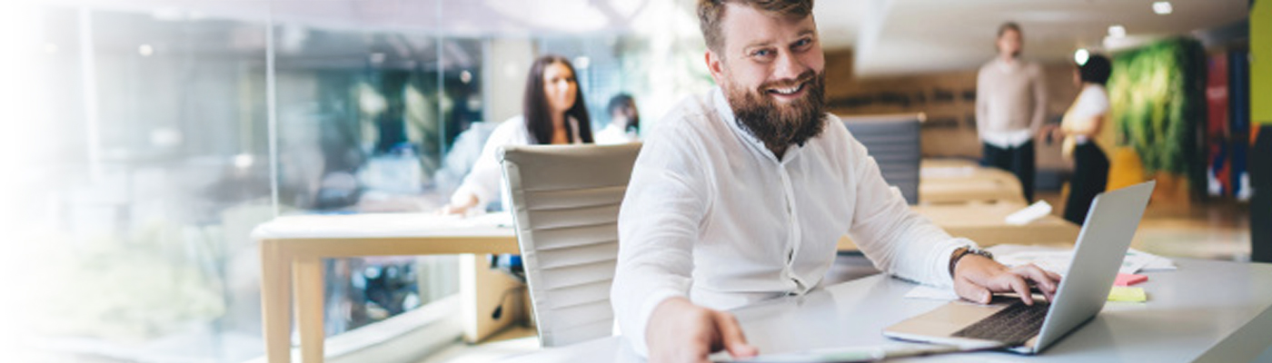 Ein junger Mann mit Bart sitzt an einem Schreibtisch mit Laptop in einem modernen Büro. Im Hintergrund sind weitere Personen sichtbar.