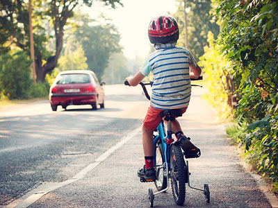 Ein Junge fährt auf einem Fahrrad entlang einer Straße, vorbeifahrende Autos und Bäume auf beiden Seiten.