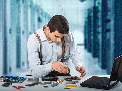 Ein Mann arbeitet an Computerhardware und verwendet Werkzeuge, während ein Laptop und verschiedene elektronische Komponenten auf dem Tisch liegen.