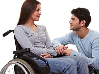 Ein Mann sitzt neben einer Frau im Rollstuhl und hält ihre Hände.