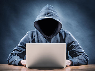 Eine Person mit Kapuze sitzt vor einem Laptop, das Gesicht ist nicht sichtbar.