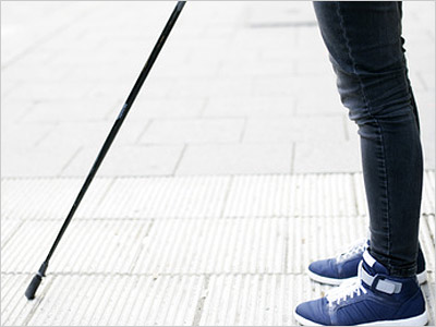 Ein Bein einer Person mit einem Stock und blauen Sneakers steht auf einem Gehweg.