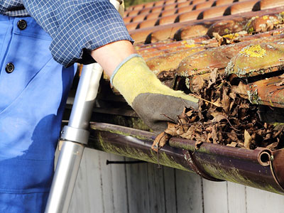 Eine Person mit Arbeitshandschuhen reinigt Laub aus einer Dachrinne.