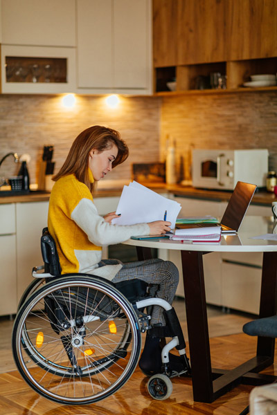 Eine Person im Rollstuhl sitzt an einem Tisch, arbeitet an einem Laptop und schaut auf Papierunterlagen.