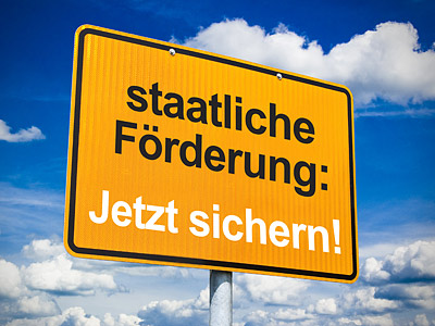 Ein orangefarbens Schild mit dem Text: "staatliche Förderung: Jetzt sichern!" vor einem blauen Himmel mit Wolken.