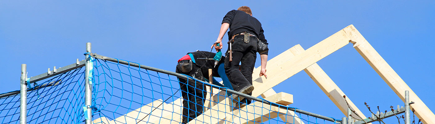 Zwei Bauarbeiter arbeiten an einem Dachgerüst unter blauem Himmel.