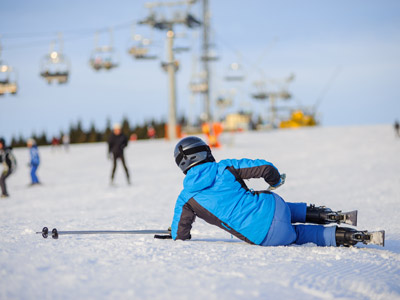 Ein Skifahrer liegt auf der Piste im Schnee, während andere Skifahrer und ein Skilift im Hintergrund zu sehen sind.