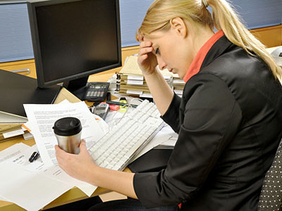 Eine gestresste Frau sitzt an einem unordentlichen Schreibtisch, hält eine Kaffeetasse und fasst sich an die Stirn.