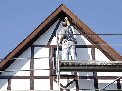 Ein Mann streicht das Dach eines Fachwerkhauses von einem Gerüst aus.