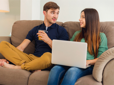 Ein Paar sitzt auf einer Couch, der Mann hält ein Getränk und die Frau arbeitet am Laptop.