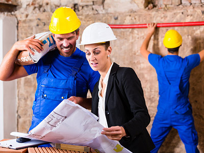 Ein Bauleiter und eine Frau schauen sich Baupläne an, während zwei Handwerker im Hintergrund arbeiten.