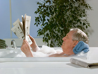Ein älterer Mann liegt entspannt in einer Badewanne mit Schaum und liest eine Zeitung.