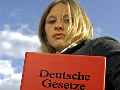 Eine Person mit langen, blonden Haaren hält ein rotes Buch mit der Aufschrift "Deutsche Gesetze" vor einem bewölkten Himmel.