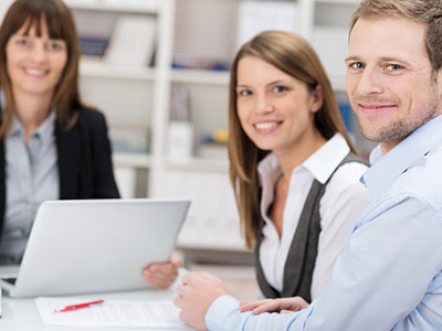 Drei Personen sitzen an einem Tisch, lächeln und arbeiten mit einem Laptop und Dokumenten.