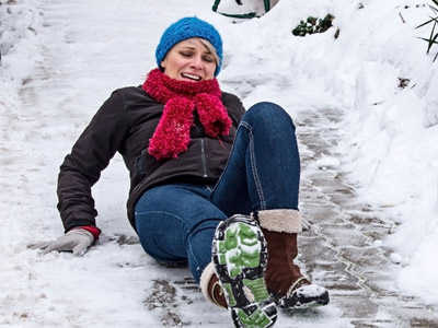 Eine Frau fällt auf einem vereisten Gehweg und hat einen verletzten Gesichtsausdruck. Sie trägt einen blauen Hut und einen roten Schal.