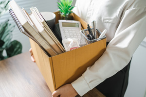 Eine Person hält einen Karton mit Büromaterialien, darunter Notizbücher, einen Taschenrechner, Stifte und eine kleine Pflanze.