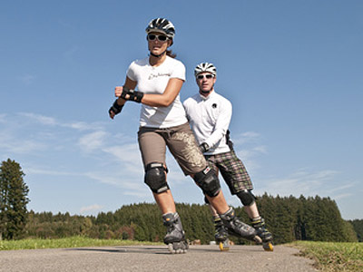 Zwei Rollschuhfahrer auf einer Strecke im Freien, umgeben von Bäumen und klarem Himmel.