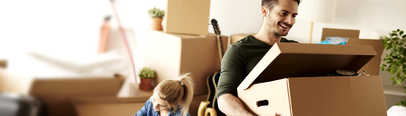 Ein Mann trägt eine Umzugskarton, während eine Frau im Hintergrund beschäftigt ist. Im Raum befinden sich weitere Umzugskartons.