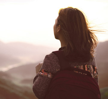 Eine Frau mit Rucksack schaut auf eine Landschaft im Hintergrund.