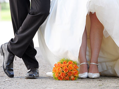 Ein Hochzeitsbild mit dem unteren Teil eines Brautpaares, das in eleganter Kleidung steht. Die Braut hält einen Blumenstrauß mit orangefarbenen Rosen.